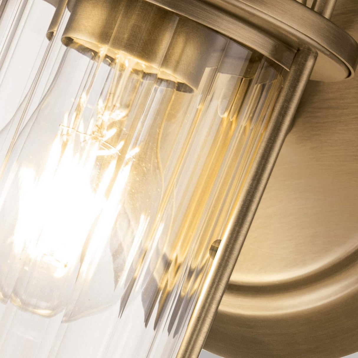 A detailed view of the Riggs 1 Light Wall Light in weathered brass, featuring an illuminated cylindrical glass shade highlights the light bulb within. This wall sconce is ideal for bathroom lighting, offering an industrial style with a sophisticated and edgy touch.