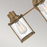 Close-up of the Riggs 3 Light Wall Light in weathered brass, featuring a clear glass enclosure that highlights an exposed filament bulb. This vintage-inspired design showcases cylindrical shapes against a neutral background, making it ideal for stylish bathroom lighting.