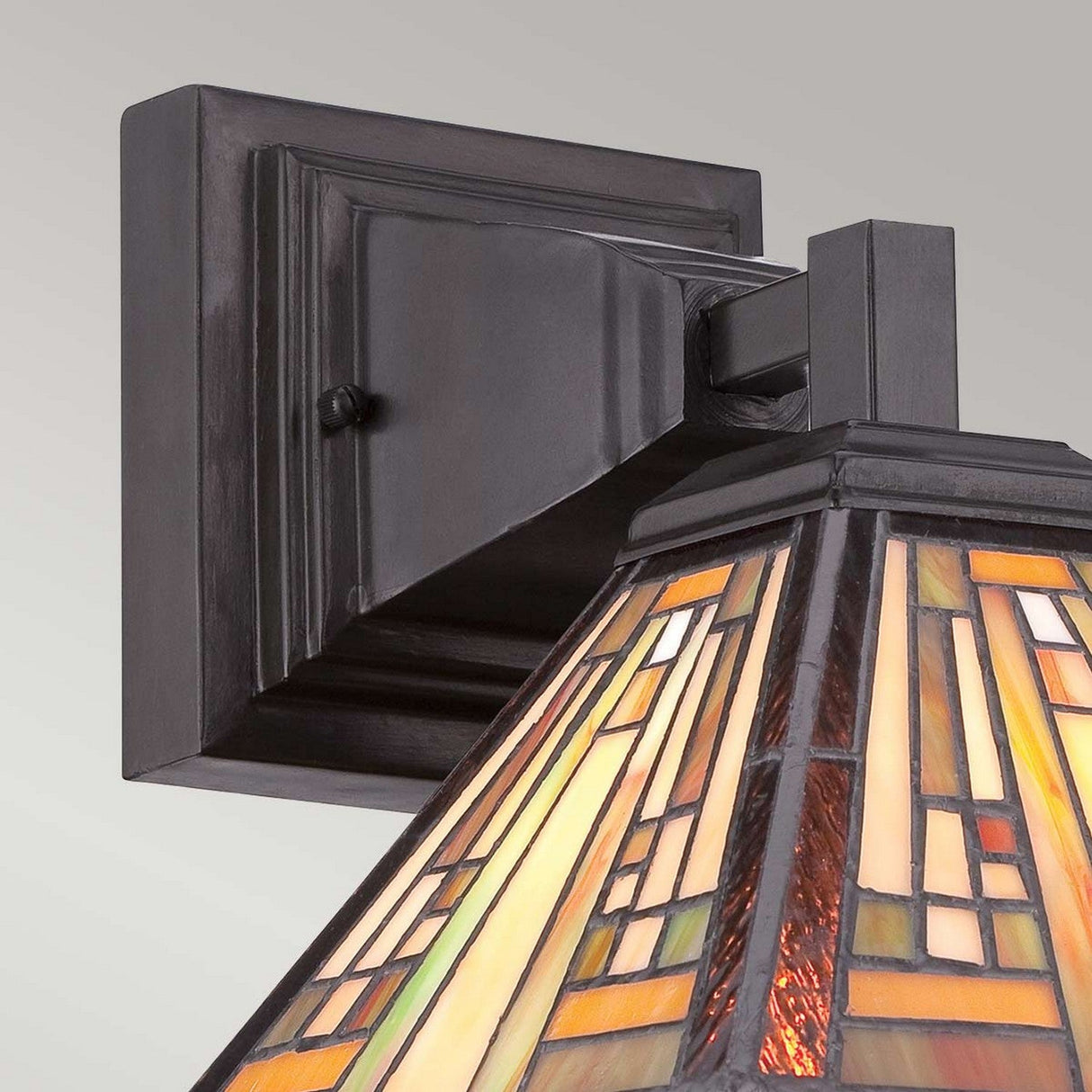 Close-up of the Stephen 1 Light Tiffany Wall Light - Vintage Bronze, showcasing a stained glass shade in amber, yellow, and cream geometric patterns. The vintage bronze mount features a dark, textured metal with a square base, offering an elegant and classic look reminiscent of the Arts & Crafts Movement.