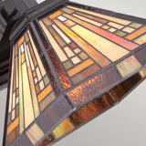 Detailed view of the Stephen 1 Light Tiffany Wall Light in a vintage bronze finish, showcasing a stained glass lampshade adorned with geometric patterns in warm shades of amber, orange, and brown. The design beautifully reflects the Arts & Crafts Movement, with its luminous glow adding to its timeless charm.