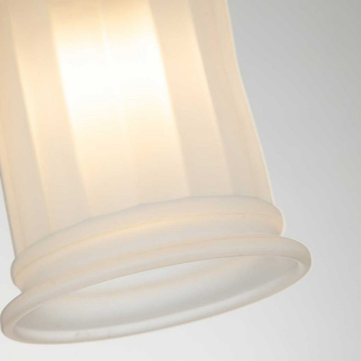 A close-up of the Swell 1 Light Wall Light with a softly glowing, frosted white glass shade. The ambient illumination creates a warm and gentle glow, highlighting the textured surface of the glass. The fixture is adorned with brushed brass, set against a soft, neutral gray background.