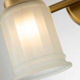Close-up of the Swell 2 Light Wall Light in brushed brass, showcasing its arm and frosted glass shade. The fixture gently diffuses light through the textured ripples of its cylindrical glass against a plain white wall.