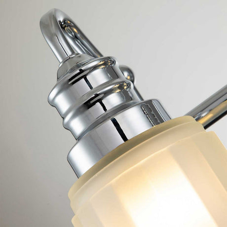 Close-up of the Swell 2 Light Wall Light in polished chrome displaying its sleek, curved design with a frosted glass shade. The light is turned on, illuminating the glass and casting a warm glow against a neutral backdrop, making it an elegant addition to the bathroom.