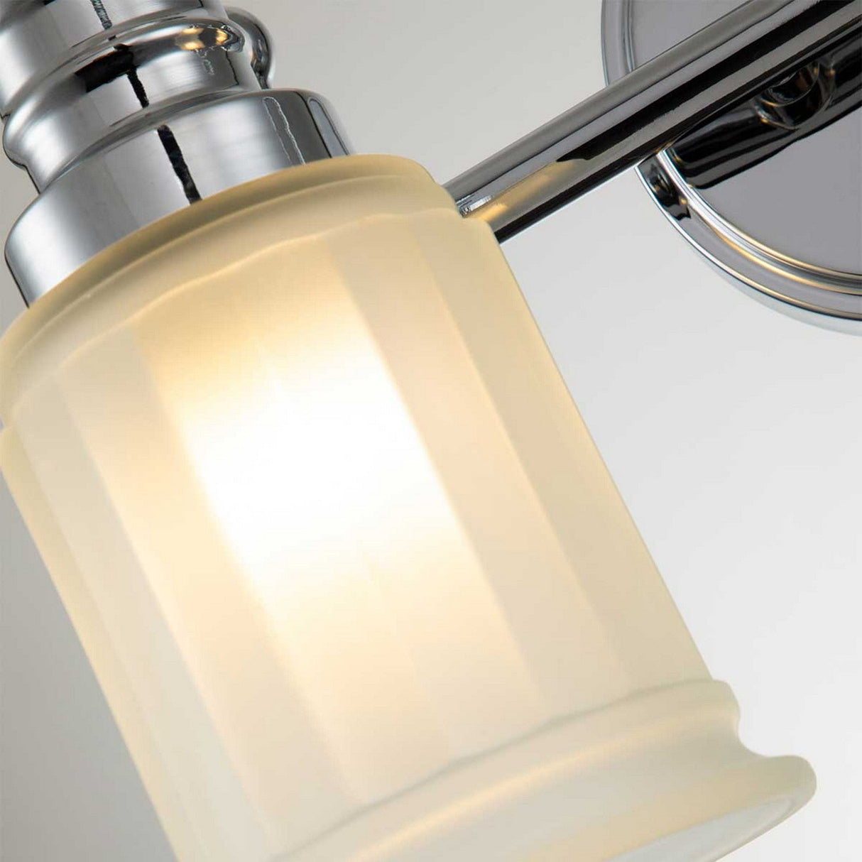 A close-up view of the Swell 2 Light Wall Light in polished chrome showcases its modern aesthetic, featuring sleek lines and a softly illuminated frosted glass shade that highlights the smooth texture of the glass and the reflective surface of the polished metal.