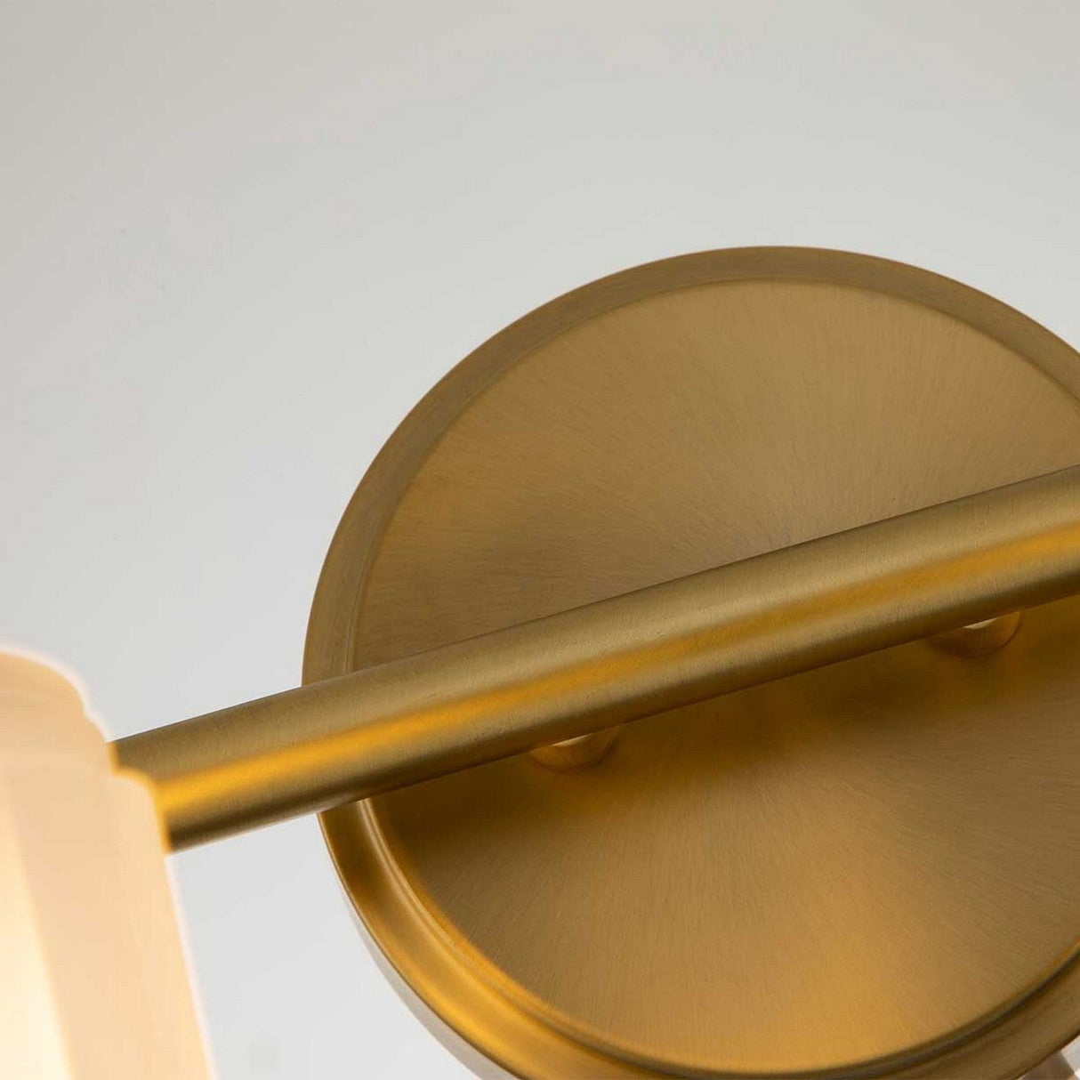 Close-up of the Swell 4 Light Wall Light in brushed brass, featuring a round, gold-colored wall mount with a horizontal cylindrical rod. This wall light has a sleek texture that casts subtle shadows, exuding metropolitan sophistication against an out-of-focus background.