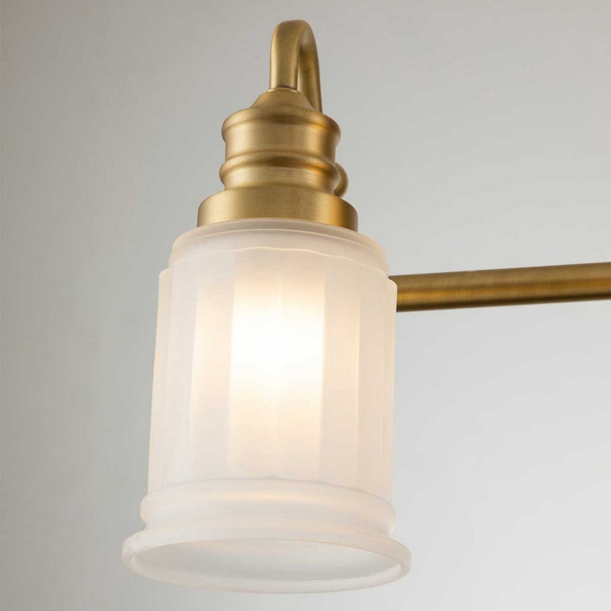 Close-up of the Swell 4 Light Wall Light in brushed brass, showcasing a vintage-style finish with a frosted glass shade and an elegant ribbed design. The softly glowing light exudes metropolitan sophistication against a neutral background.