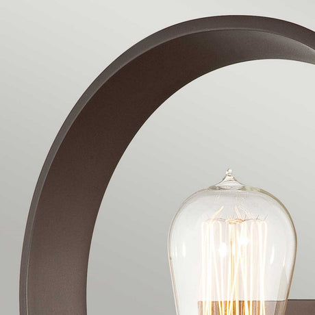 A close-up of the Theatre Row 1 Light Wall Light in Western Bronze features a cylindrical glass bulb, highlighting the exposed bulb design. It is encased in a sleek, curved metal frame set against a neutral backdrop, embodying a minimalist and contemporary style.