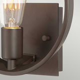 A close-up of the Theatre Row 1 Light Wall Light in Western Bronze reveals a modern wall sconce with a black metal frame. This design showcases an exposed bulb style with a glowing filament, evocative of vintage theatre lighting, set against a light gray background.