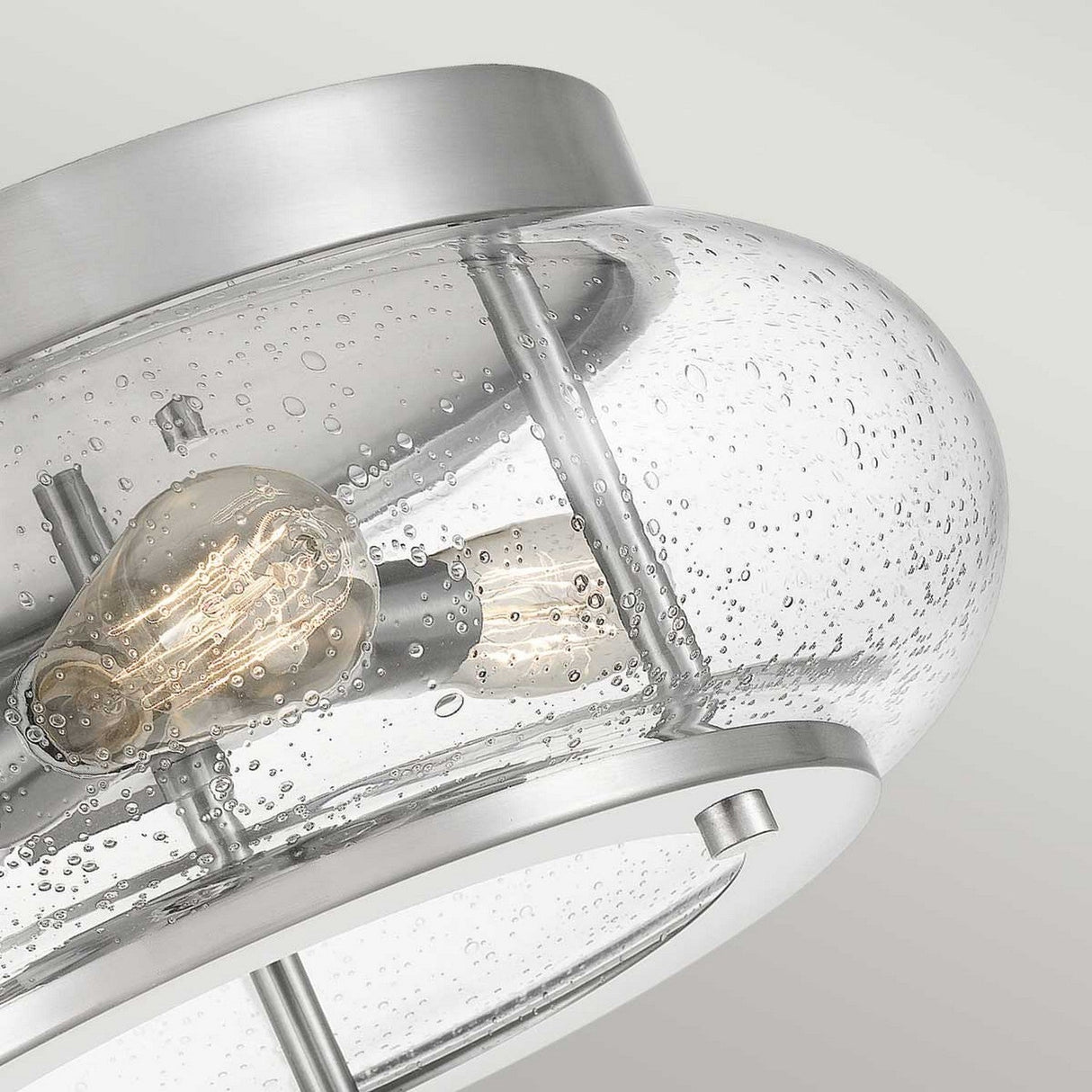 A close-up of the Trilogy 3 Light Flush showcases a modern ceiling light fixture with a clear, seeded glass shade adorned with tiny bubbles. Inside, you can see two Edison-style LED bulbs, while the brushed nickel frame adds to its contemporary appearance.