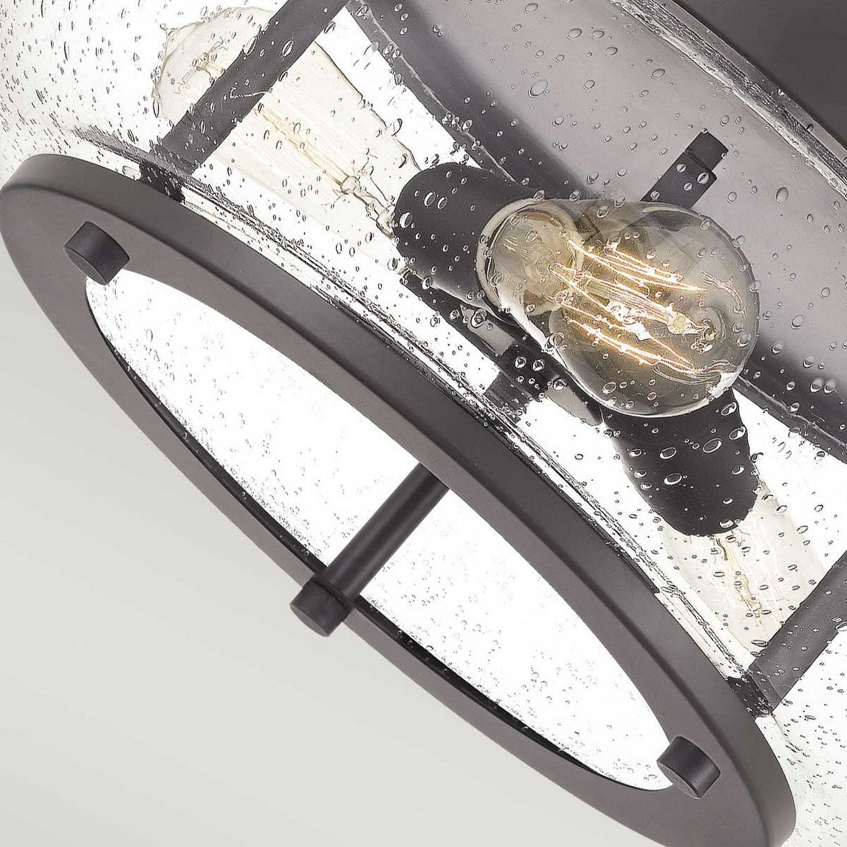 A close-up of the Trilogy 3 Light Flush, showcasing its modern design with a clear seeded glass shade and an Edison-style LED filament bulb. The fixture is crafted with an Old Bronze metal frame and embodies a minimalist aesthetic, enhancing its contemporary appeal.