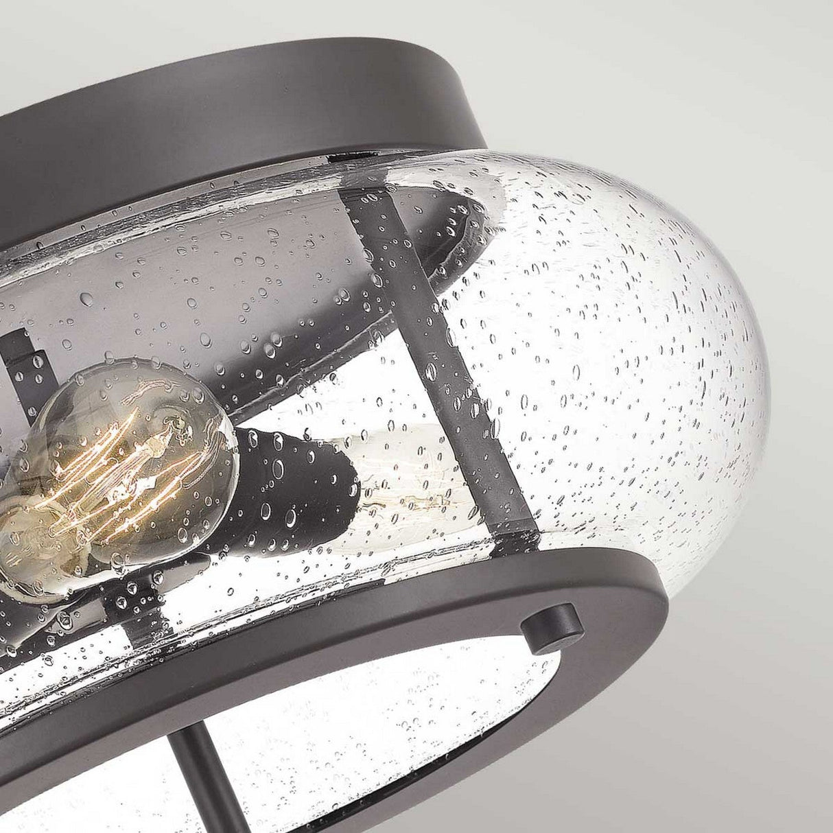 Close-up of the Trilogy 3 Light Flush featuring a clear, seeded glass dome and an Edison-style LED filament bulb. The fixture's old bronze metal frame adds vintage charm against the light gray background.