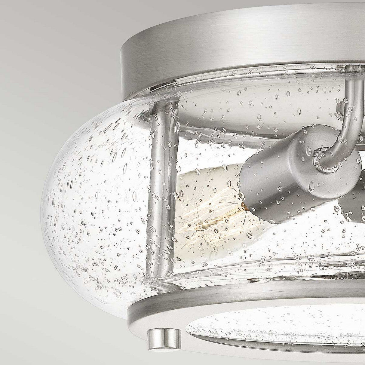 Close-up of the Trilogy 2 Light Flush in a brushed nickel finish. It includes a clear, seeded glass shade with a bubbled texture, highlighting an exposed light bulb within. The modern and minimalist design is set against a neutral background.