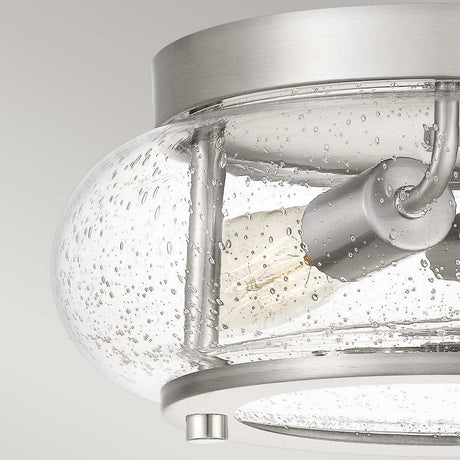 Close-up of the Trilogy 2 Light Flush in a brushed nickel finish. It includes a clear, seeded glass shade with a bubbled texture, highlighting an exposed light bulb within. The modern and minimalist design is set against a neutral background.