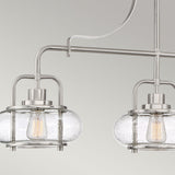 Two Trilogy 3 Light Island Lights in brushed nickel, featuring clear seeded glass shades and exposed filament bulbs, hang from a sleek bar. The elegant design contrasts with a simple light gray background.