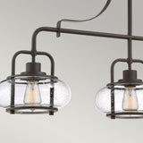 A close-up of the Trilogy 3 Light Island Light in a dark, brushed Olde Bronze finish features two hanging glass globe lamps showcasing LED filament bulbs. The textured, bubbled glass enhances its vintage charm against a neutral, light background.