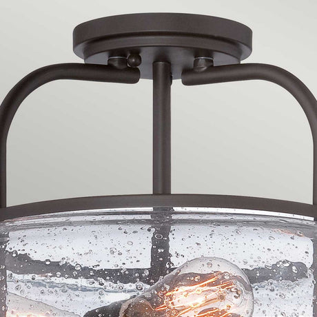 Close-up of the Trilogy 3 Light Medium Semi-Flush fixture in an Old Bronze finish, featuring a textured seeded glass cover. Inside, an incandescent bulb glows warmly, casting light against a light gray background and capturing the essence of industrial lighting.