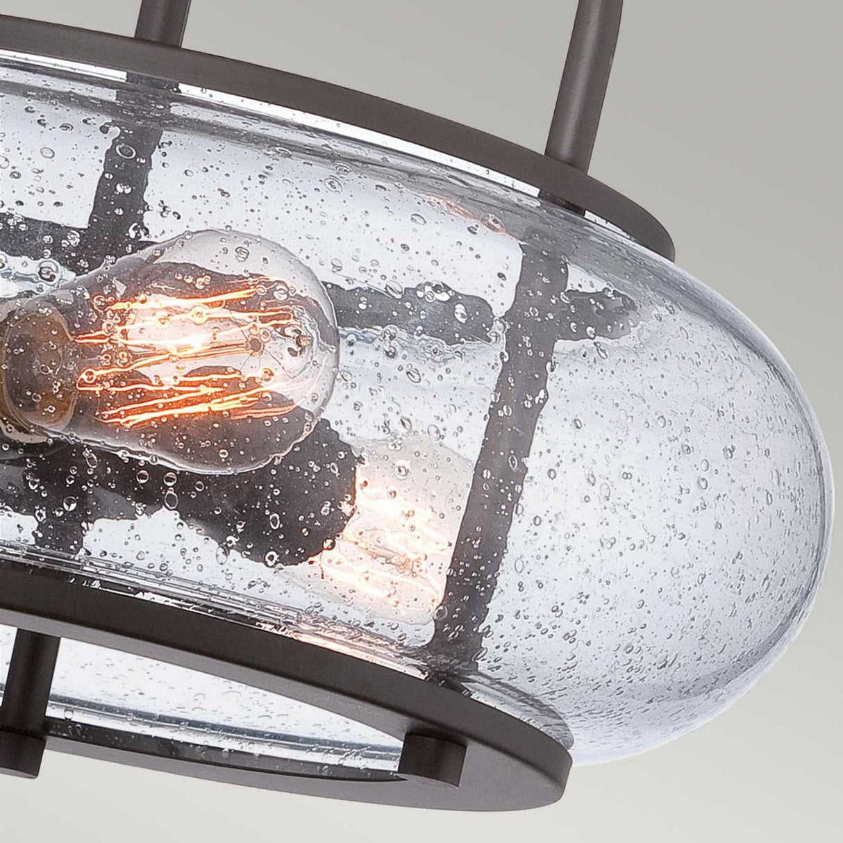 Close-up of the Trilogy 3 Light Medium Semi-Flush in Old Bronze, showcasing an Edison bulb inside clear, seeded glass. Its dark metal frame highlights a vintage yet contemporary design against a light gray background, seamlessly aligning with industrial lighting aesthetics.