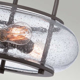 Close-up of the Trilogy 3 Light Medium Semi-Flush in Old Bronze, showcasing an Edison bulb inside clear, seeded glass. Its dark metal frame highlights a vintage yet contemporary design against a light gray background, seamlessly aligning with industrial lighting aesthetics.