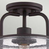 Close-up of the Trilogy 1 Light Small Semi-Flush in Old Bronze, featuring a dark metal frame and a seeded glass shade. This fixture embodies a modern, industrial design with an Edison-style LED filament bulb visible through the textured glass.