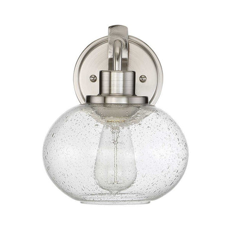 The Trilogy 1 Light Wall Light - Brushed Nickel showcases a clear glass globe with a distinctive seeded texture, elegantly displaying the visible bulb. Its design masterfully combines modern and vintage elements for a timeless allure.