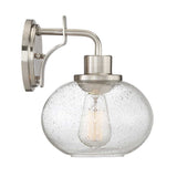 The Trilogy 1 Light Wall Light in brushed nickel features a clear, seeded glass globe with a textured, bubble-like pattern that encases an exposed vintage-style bulb, offering a modern and elegant design.