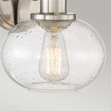 A close-up of the Trilogy 1 Light Wall Light - Brushed Nickel showcases a wall-mounted fixture with a clear seeded glass globe featuring bubble patterns. Inside, a vintage-style filament bulb emits a warm glow. The fixture has a brushed nickel finish and is set against a light gray background.