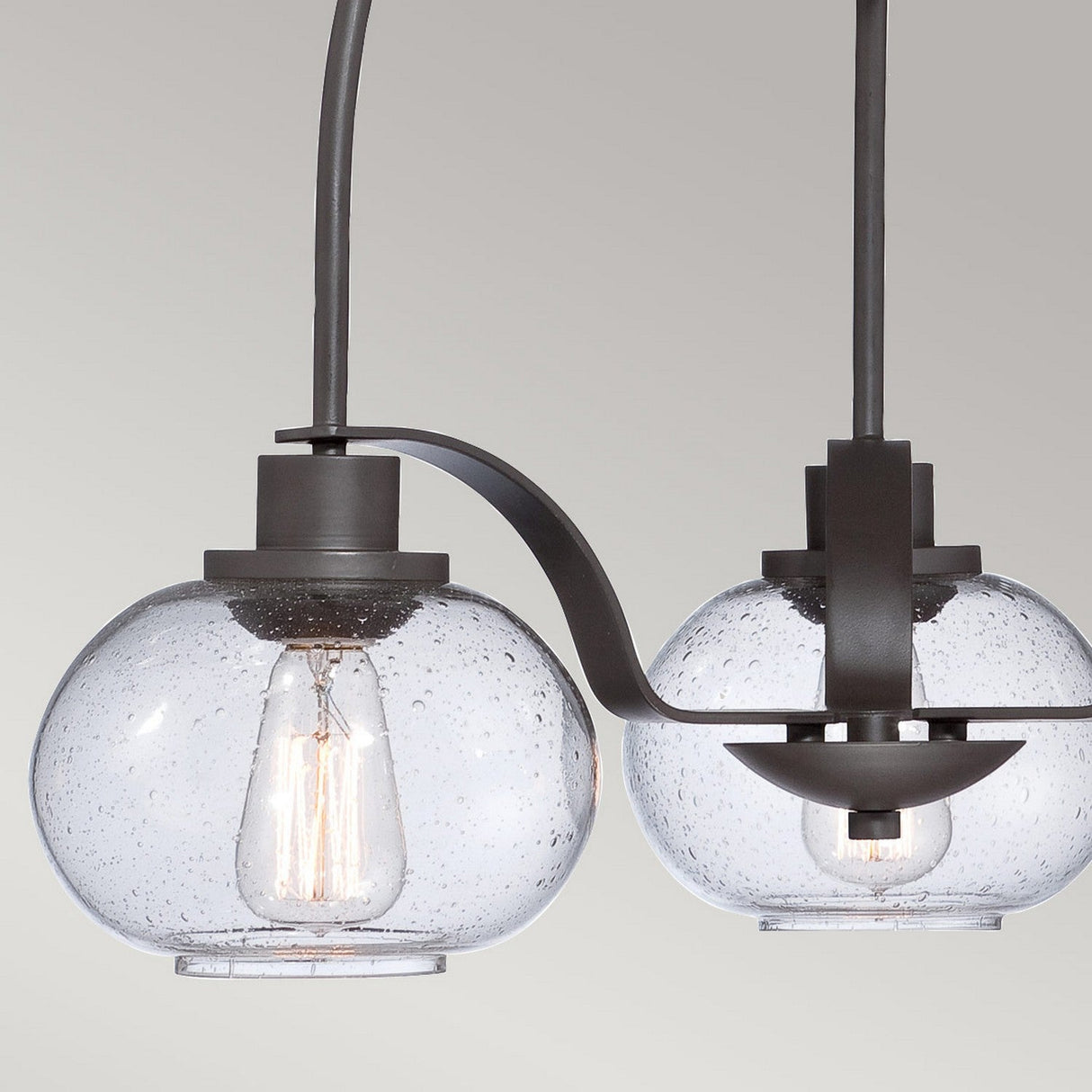 A detailed view of a contemporary light fixture featuring two clear, seeded glass globes that reveal visible filaments. The fixture is elegantly finished in Old Bronze with a dark metal frame and curved design, offering a striking resemblance to the Trilogy 3 Light Chandelier - Old Bronze, set against a neutral backdrop.