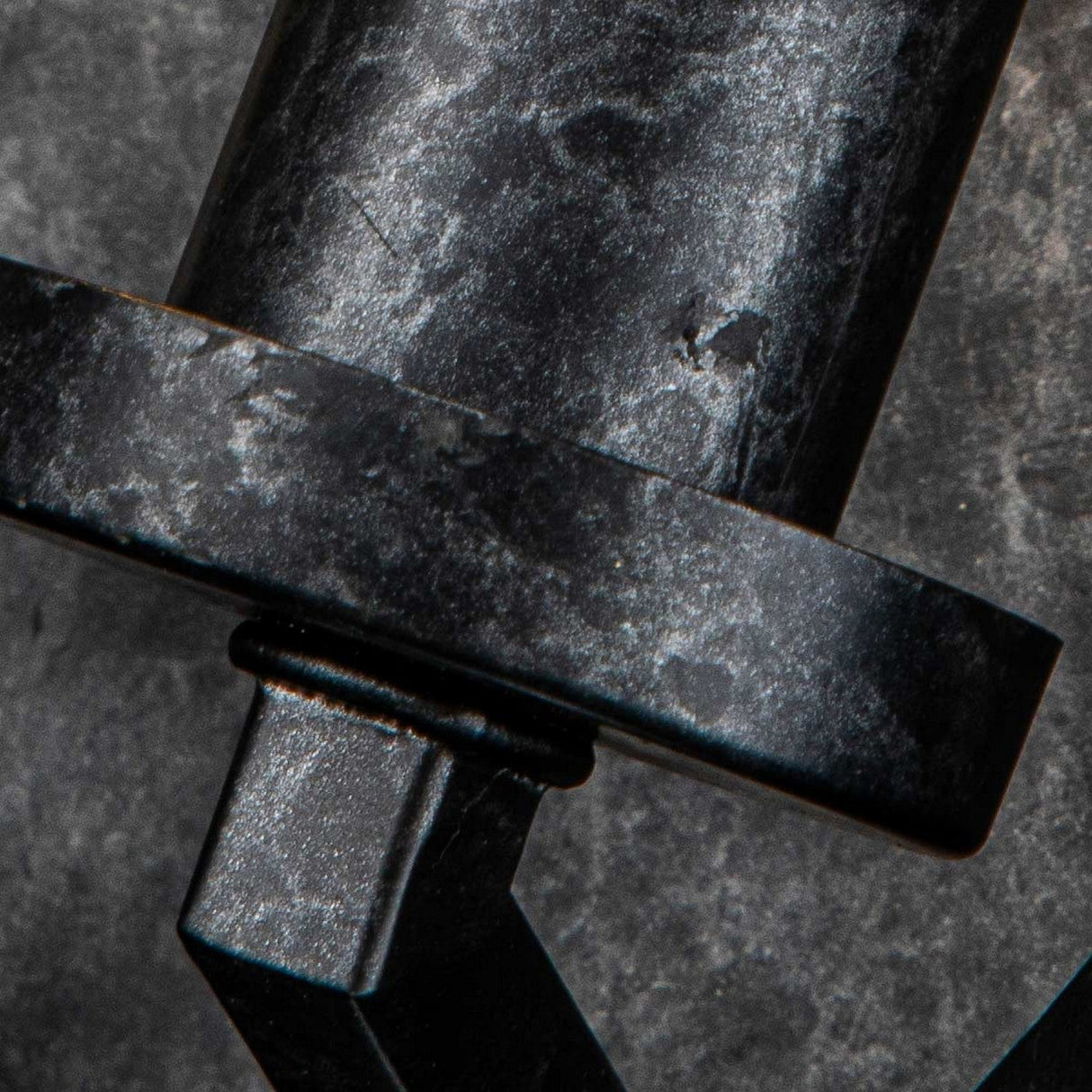 A close-up view of the Vanguard 2 Light Wall Light in Old Black reveals a weathered, cylindrical metal object with a flat circular component. Its surface exhibits an industrial-inspired design, showcasing signs of wear and texture that lend it a rugged appearance against a mottled background.