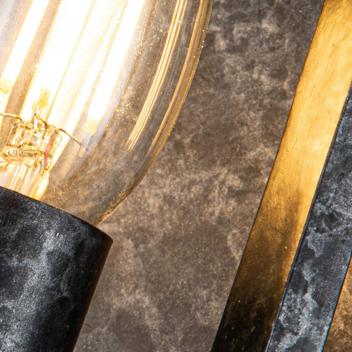 Close-up of the Vanguard 2 Light Wall Light - Old Black, featuring a glowing bulb with a visible filament. The industrial-inspired design is finished in Old Black and stands out against a textured gray surface, highlighting the warm light it emits.