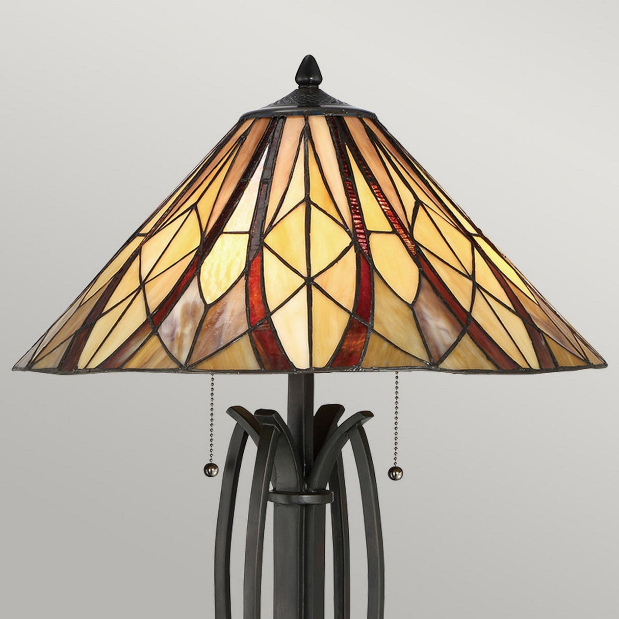 The Victory 2 Light Tiffany Table Lamp - Valiant Bronze showcases a decorative geometric pattern in shades of amber, brown, and cream. It includes two pull chains and boasts a base with elegant curved details finished in valiant bronze.