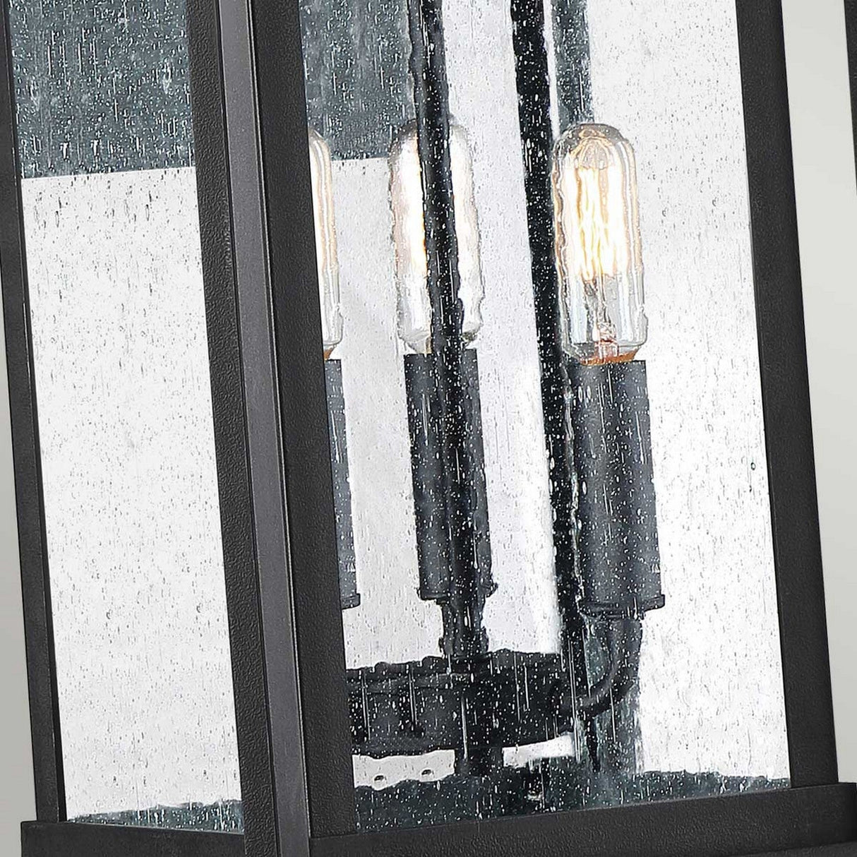 Here's a rewritten version of your sentence:

Close-up of the Wakefield Outdoor 3 Light Large Wall Lantern in Earth Black, featuring a metal frame that encases vertical, exposed filament bulbs. The glass is textured with raindrops, creating a diffused glow that embodies the classic Arts and Crafts lighting style.