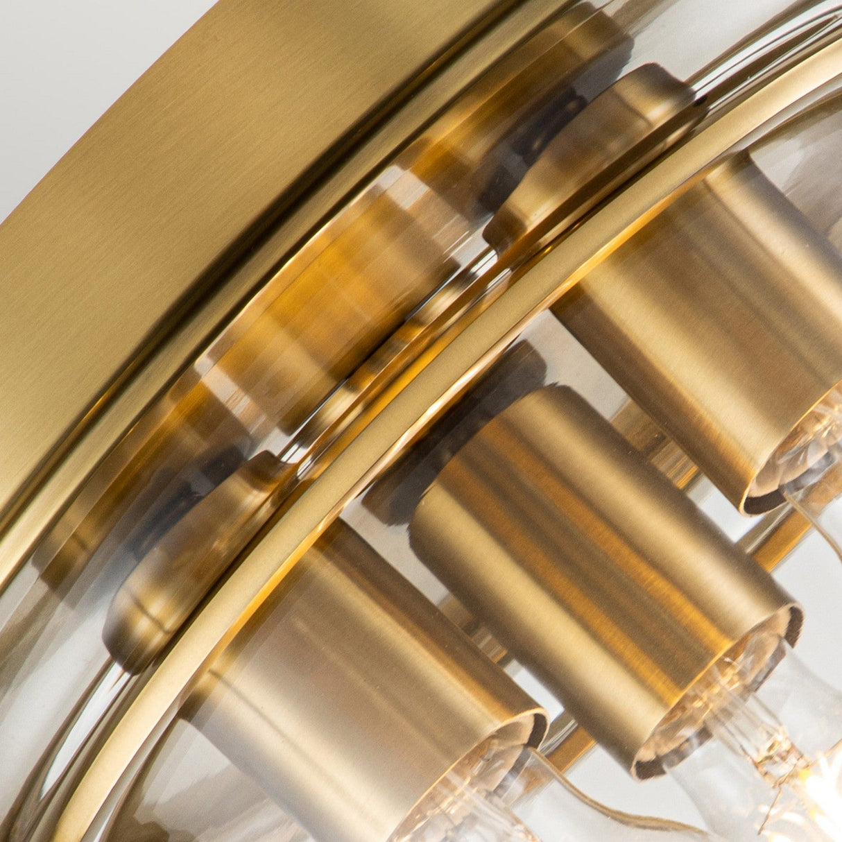Close-up of the Whistling 3 Light Flush in brushed brass, featuring visible bulbs. The design includes cylindrical brass holders and a curved glass cover, presenting a modern and elegant aesthetic. Ideal for bathroom lighting with its IP44 rating.