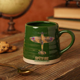 Green ribbed stoneware mug printed with a camouflage Spitfire plane and SPITFIRE text, unglazed base band, shown on a wooden table with books and a spoon.