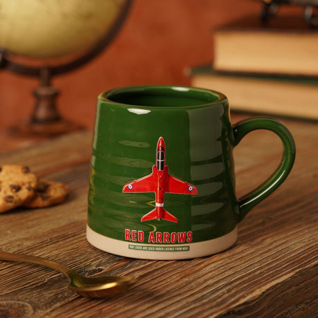 Green ribbed stoneware mug with beige base featuring a red Red Arrows jet and bold "Red Arrows" text, shown on a wooden table with books, a globe, and cookies.