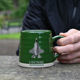 Green ribbed stoneware mug with beige base, held by hand outdoors, featuring a grey F-35B Lightning jet design and "LIGHTNING" text beneath it.