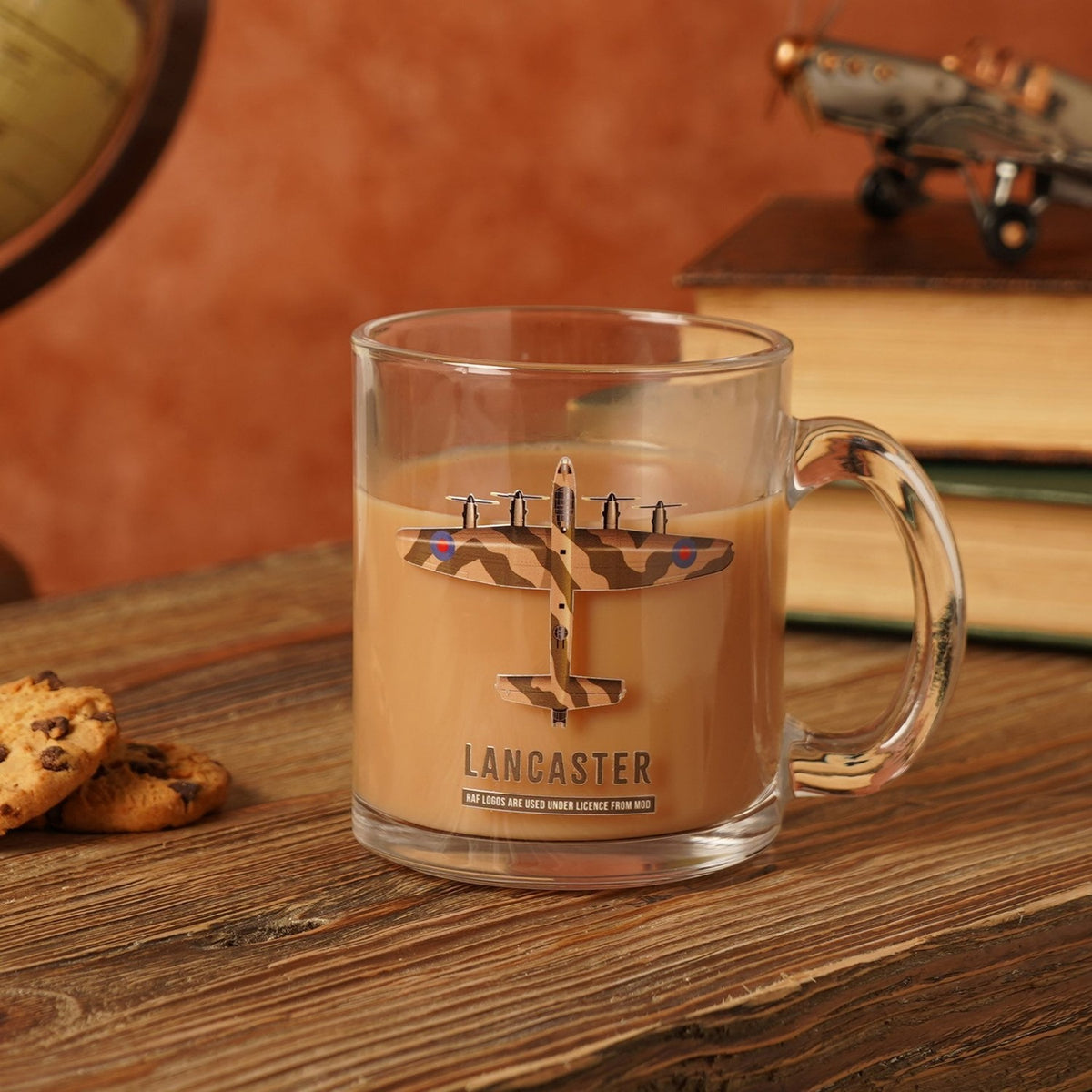 Clear glass mug filled with coffee, printed with a camouflage Lancaster bomber design and "LANCASTER" text, shown on a wooden surface with books and cookies.