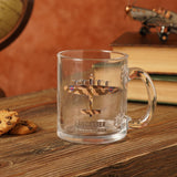 Clear glass 10oz mug with a camouflage print Lancaster bomber design, showing brown and tan tones, wing roundels, and "LANCASTER" text, displayed on a wooden surface with books and a model plane.
