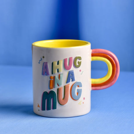 White ceramic mug with raised 3D multicoloured "A Hug in a Mug" lettering, yellow glazed interior, and a layered pink and yellow rainbow-shaped handle, on a blue background.