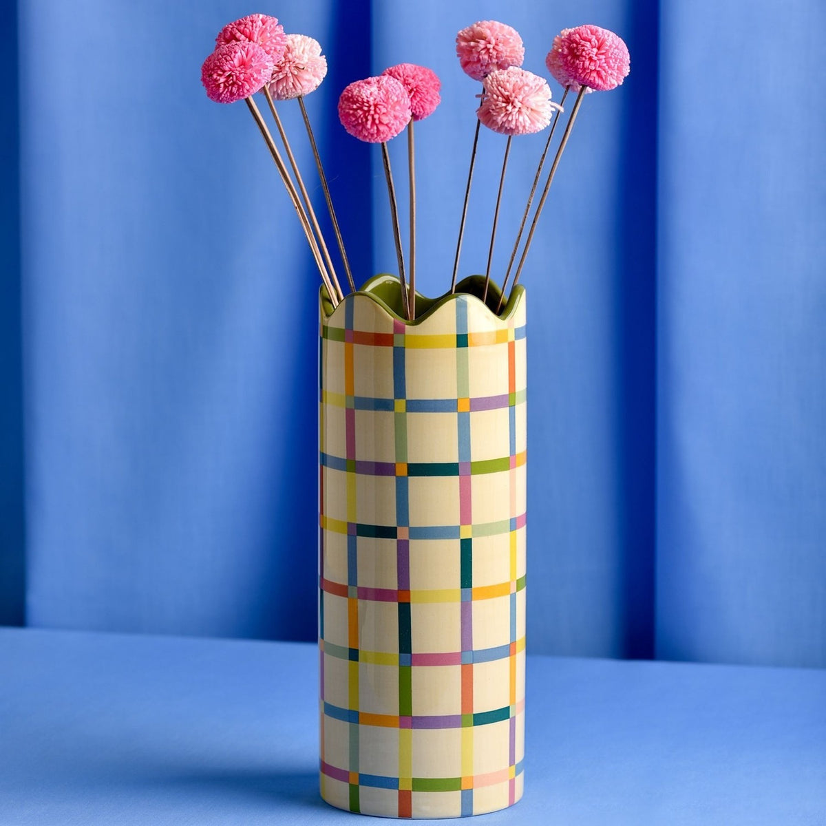 Cylindrical ceramic vase, 25.3cm tall, with a multicoloured check pattern on a yellow ground and a green wavy rim, shown with pink pom-pom stems against a blue background.