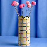 Cylindrical ceramic vase, 25.3cm tall, with a multicoloured check pattern on a yellow ground and a green wavy rim, shown with pink pom-pom stems against a blue background.