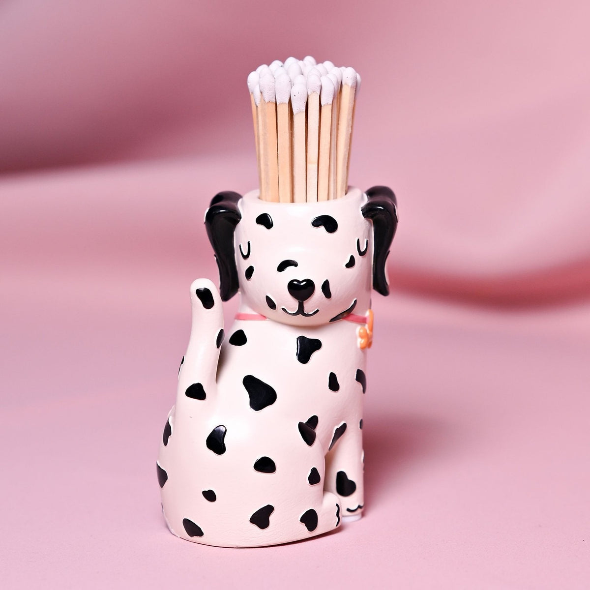 Spotted Dog Resin Matchstick Holder in white, sitting with black-spotted markings, floppy ears, pink collar, and matches in its head opening, set against a pink background.