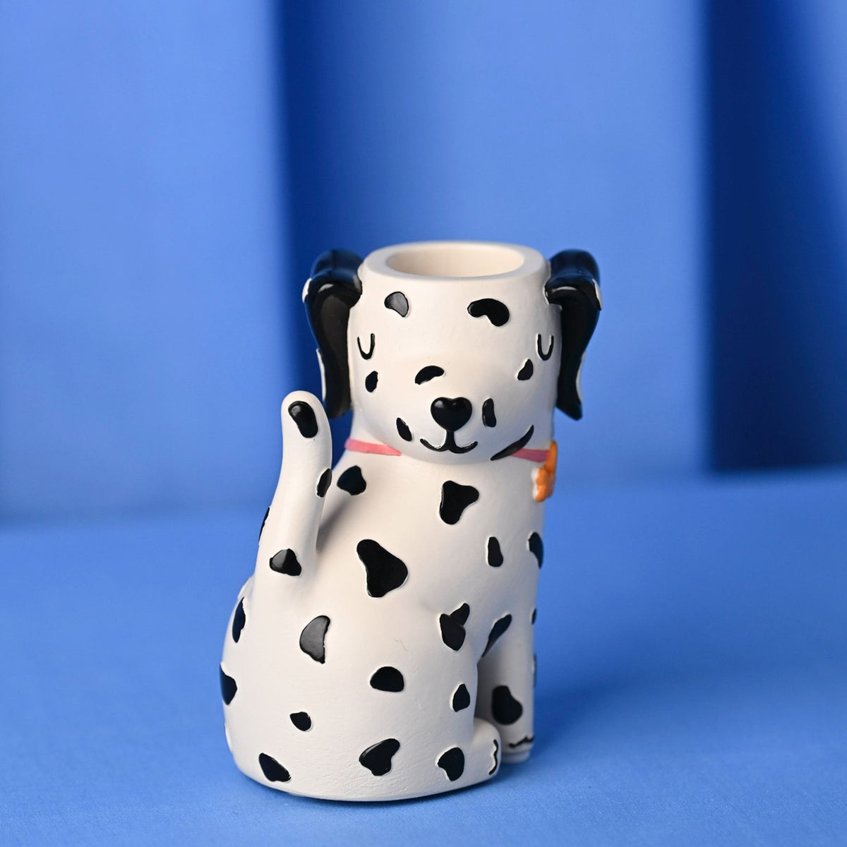 White resin matchstick holder shaped as a seated spotted dog with black ears and spots, pink collar, curled tail, and a matchstick opening atop its head, on a blue background.