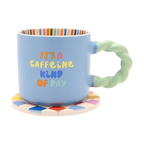 Pale blue porcelain mug with twisted mint-green handle and "It's A Caffeine Kind Of Day" text, rainbow-striped interior, set on a round diamond-pattern coaster against a white background.