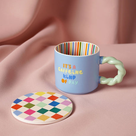 Blue porcelain mug with twisted mint-green handle, colourful "It's A Caffeine Kind Of Day" text, striped interior, and matching round checkered coaster on a pink fabric backdrop.