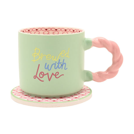 Green porcelain mug with twisted pink handle, "Brewed with Love" in yellow, blue and pink script, on a matching patterned round coaster against a white background.