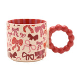 Porcelain mug with cream body patterned in red, pink, and burgundy bows, featuring a chunky red bobble-ring handle and red striped interior, shown on a plain white background.