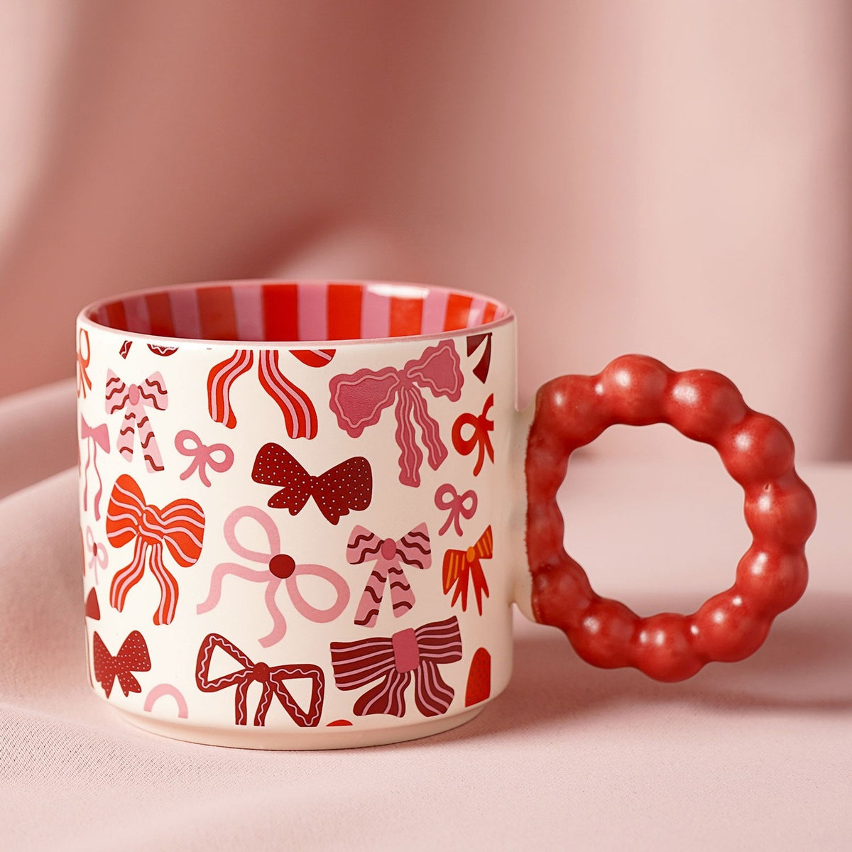 Porcelain mug with cream body printed with red, pink, and burgundy bow patterns, featuring a chunky red bobble-shaped handle and red and pink striped interior, set against a pink background.