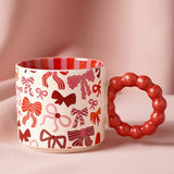 Porcelain mug with cream body printed with red, pink, and burgundy bow patterns, featuring a chunky red bobble-shaped handle and red and pink striped interior, set against a pink background.