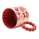 Porcelain mug lying on its side showing a red striped interior and a chunky bobble-style red ring handle, with a cream exterior patterned in pink and burgundy bows, on a plain white background.