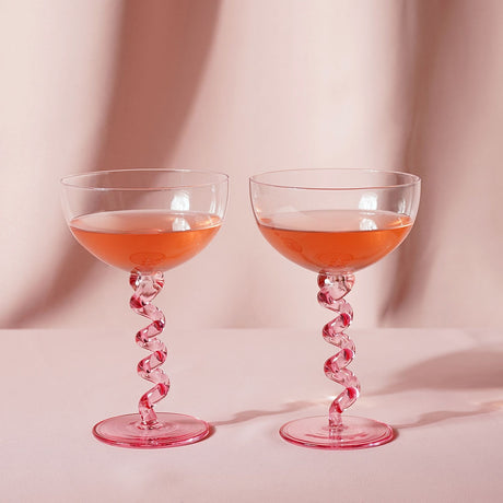 Pair of 300ml coupe glasses with clear wide bowls on pink wiggle stems and round pink feet, filled with orange liquid against a soft pink backdrop.